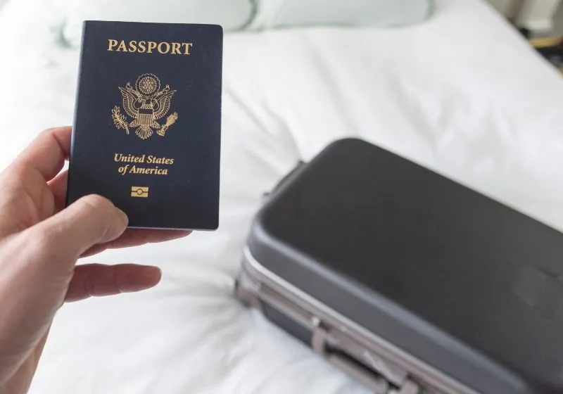 mão de uma pessoa segurando um passport e atrás uma mala em cima de uma cama 