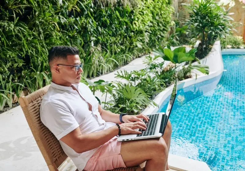 homem sentado em uma cadeira mexendo em um computador enfrente uma piscina e atrás várias plantas