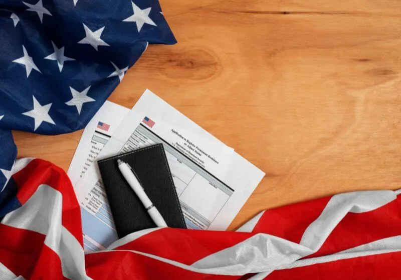 imagens de uma caneta com um mini caderno e folhas em baixo e do lado a bandeira dos Estados Unidos em um fundo de madeira