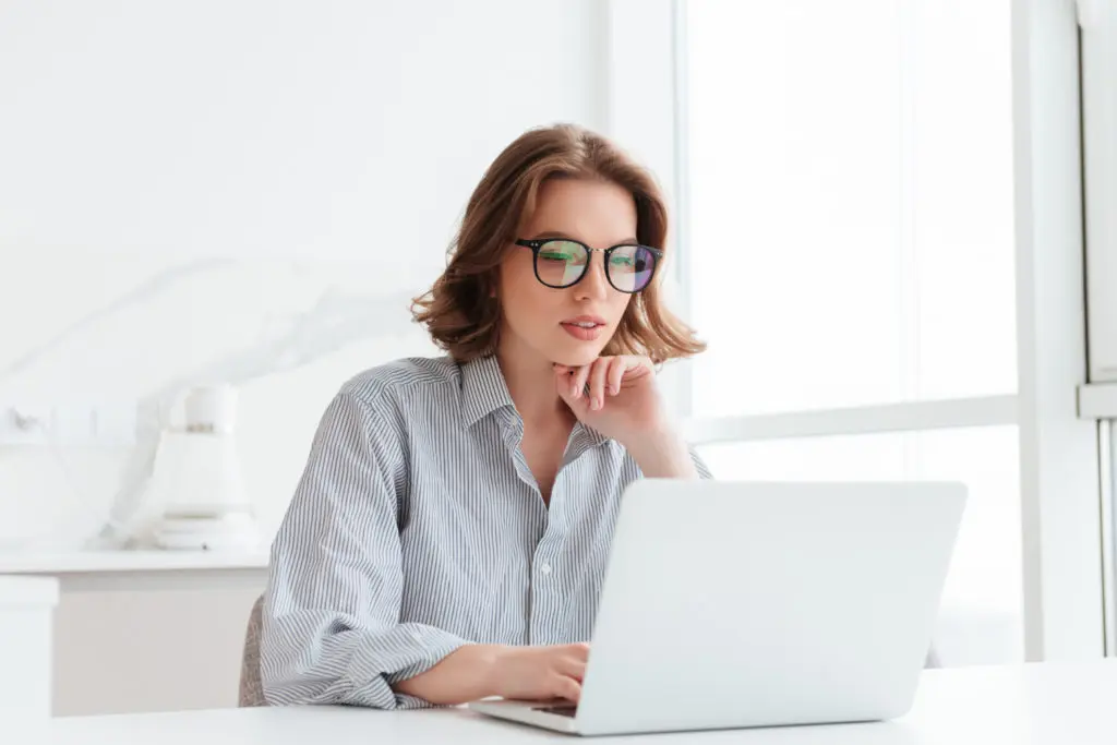 charmosa-empresaria-de-oculos-e-camisa-listrada-trabalhando-com-o-computador-portatil-enquanto-localizacao-em-casa Imagem de mulher branca de óculos usando notebook para fazer revisão de texto.